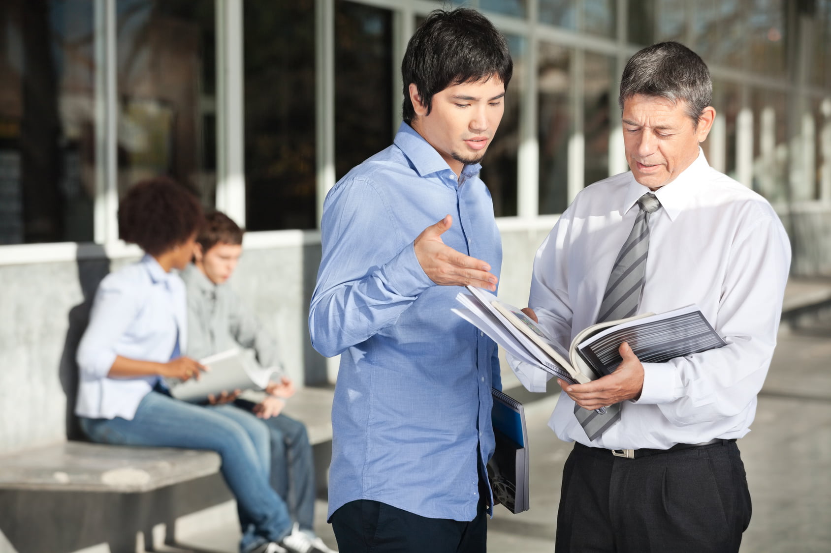 Mature male teacher holding books while discussing with student on college campus